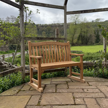 Load image into Gallery viewer, Oxford 1.2m memorial bench with optional set of feet on a stone patio with a view of green fields and trees.