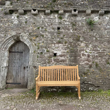 Load image into Gallery viewer, FSC certified Teak Warwick memorial bench with a set of feet in front of stone castle walls