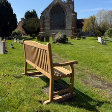 Load image into Gallery viewer, Teak Warwick memorial bench with a set of feet in a cemetery with a church in the background